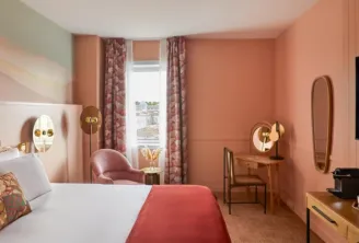 Elegant hotel bedroom with coral walls, white bed with gold palm-shaped headboard, pink armchair, floral curtains, wooden desk and wall-mounted television.