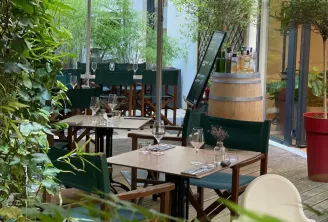 Shaded outdoor dining terrace at Les Tables Vatel restaurant in Bordeaux featuring wooden tables, beige umbrellas, lush greenery, and a wine barrel in the background