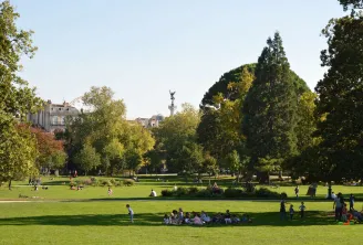View of Bordeaux Public Garden featuring lush green lawns, tall trees, relaxing visitors, a monument with statue in the background and historic buildings under clear sky.