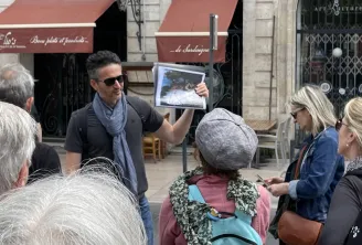 A tour guide shows a tablet to a group of visitors on a Bordeaux street, in front of historic buildings and burgundy awnings.