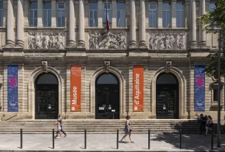Facade of the Museum of Aquitaine in Bordeaux featuring tall columns, sculptural friezes, orange and blue banners, three arched doors, and visitors in the square.