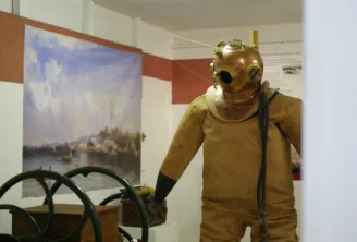 Vintage diving suit mannequin in yellow wetsuit and gilded bronze helmet displayed in a museum, with a ship's wheel and river landscape painting visible behind it.