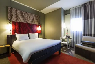 Modern hotel room featuring a double bed with white linens, red and brown headboard, pale yellow walls, warm ambient lighting, and a window with gray curtains at Ibis Bordeaux Centre Gare Saint-Jean.
