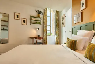 Modern and elegant hotel bedroom featuring a queen-size bed with green headboard, yellow striped cushions, tall window with gold curtains, wooden desk, round mirror, green wall shelves and contemporary décor in Bordeaux