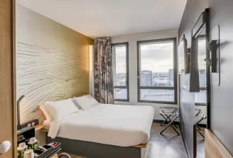 Modern hotel room with double bed with white linens, yellow headboard with wavy patterns, large windows overlooking Bordeaux cityscape, contemporary furnishings and view of the Bassins-à-Flot.