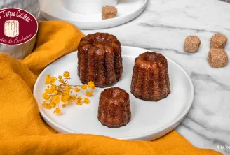 Three golden and shiny Bordeaux canelés presented on a white plate, decorated with yellow dried flowers, with orange fabric and crystallized sugars in the background
