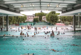 Municipal swimming pool Judaïque Jean Boiteux in Bordeaux featuring indoor pools with retractable roof, crowds of swimmers and sunbathers, exterior patio terrace, and historic buildings visible in the background