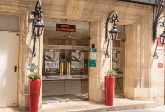 Entrance facade of Hôtel Sainte Catherine, a Quality Hotel in Bordeaux, featuring glass doors, wrought iron wall lanterns, and red welcome planters