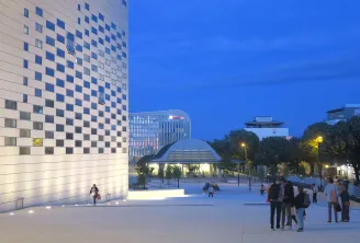 Night view of La Méca in Bordeaux featuring its illuminated white checkered facade, a paved public plaza with visitors, and modern buildings against a twilight sky.