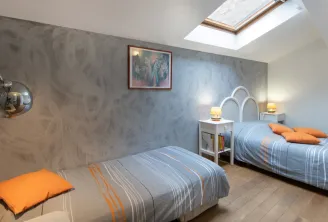 Guest bedroom in Bordeaux featuring two twin beds with blue and orange striped linens, chrome arc lamp, light gray walls, skylight, white bedside tables, and minimalist decor.