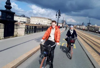 Two smiling cyclists riding bikes on a dedicated bike lane crossing a historic bridge in Bordeaux, with classical buildings and a cathedral in the background