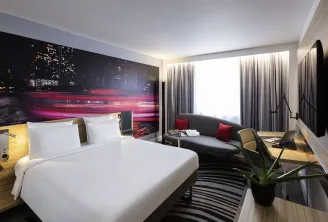 Modern hotel room at Novotel Bordeaux Centre featuring a king-size bed with white linens, a night cityscape wall mural in red and gold tones, grey sofa with red cushions, floor-to-ceiling grey curtains, and contemporary amenities.