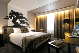 Modern hotel room featuring a queen-size bed, black dragon wall art, golden curtains, desk with laptop, and contemporary black and gold furnishings.