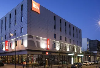 Modern facade of Ibis Bordeaux Centre Bastide hotel at night, featuring contemporary gray and white architecture, illuminated red signage, and parked cars in front