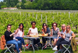 Seven women seated around a table with wine glasses and charcuterie, in a Bordeaux vineyard with buildings and trees in the background