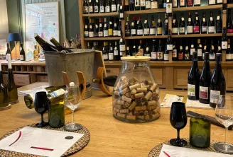 Interior of a Bordeaux wine cellar with a tasting counter displaying bottles, glasses, an ice bucket, and a jar filled with wine corks, facing shelves stocked with wines.