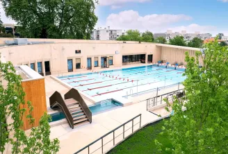 Aerial view of Jean Zay Swimming Pool in Bordeaux, featuring an Olympic-size swimming basin with marked lanes, a shallow learning pool, access stairs, beige administrative building, and surrounding greenery.