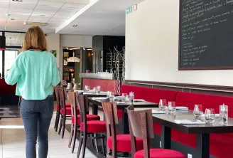Interior of a Bordeaux restaurant showing a woman in a turquoise shirt standing before a row of tables with red banquettes, wooden chairs, water glasses, and a chalk menu board on the wall.