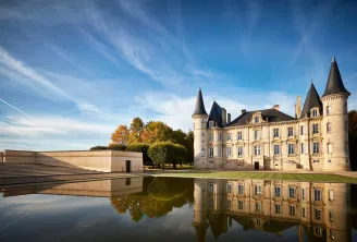 Château Pichon Baron in Bordeaux featuring distinctive conical towers and stone façades, reflected in still water, surrounded by manicured gardens and outbuildings