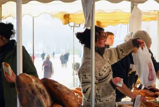 Baker displaying artisanal bread under a market tent, serving customers selecting fresh bakery products.