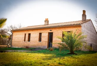 Small wooden cottage with honey-colored walls, tiled roof, stone chimneys, rectangular windows and black entrance door, surrounded by a grassy garden and tropical plants.