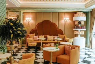 Elegant Art Deco lounge featuring ochre orange furniture, black and white checkered flooring, spherical golden chandelier, sage green and beige walls, yellow floral arrangement and green plants.