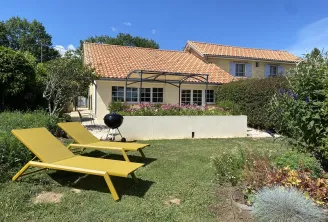 Sunny garden view of Château La Gontrie featuring a yellow lounger, manicured lawn, paved terrace with BBQ, and tile-roofed house in the background.