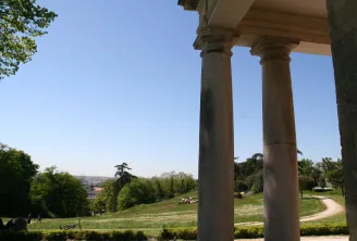 View from a stone colonnade at Palmer Park in Bordeaux, overlooking green lawns, trees, and the city skyline in the distance under clear blue sky.
