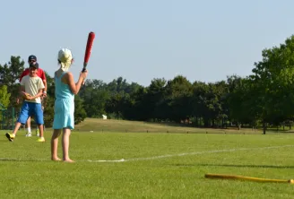 Child holding a red baseball bat on a grass sports field at Domaine du Pinsan in Bordeaux, with adults supervising and other children in the background.