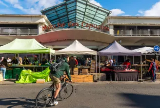 View of the Marché des Capucins in Bordeaux with colorful market stalls, vendors and customers, a cyclist passing by, and the main entrance with its glass canopy in the background.