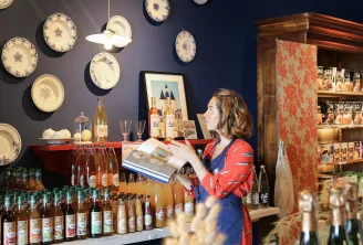 A saleswoman displays products at the Echoppe de la Lune in Bordeaux, surrounded by wine and spirits bottles, with decorative blue and white plates hanging on the wall.