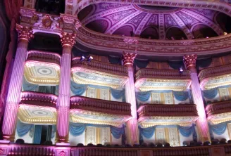 Interior view of the Grand Théâtre of Bordeaux lit in pink, displaying ornate boxes with Corinthian columns, blue and gold draperies, and the architectural dome of the ceiling.