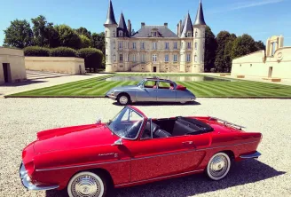 Two classic cars displayed in front of a French château with turrets: a grey Citroën DS in the background and a red convertible from the 1950s in the foreground, parked on a gravel driveway with striped lawn.