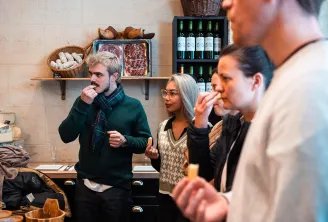 Food tour guide presenting cured meats and wines to visitors in Bordeaux, standing in front of shelves displaying Iberian ham, bread, and bottles of red wine.