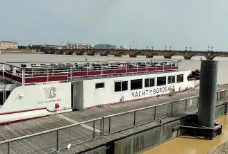 The Maddalena, a white and red river cruise yacht, moored at a Bordeaux quay with the Garonne river and Pont de Pierre bridge visible in the background.