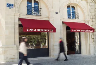 Storefront of a wines and spirits shop 'La Cave du Château' in Bordeaux, located on Place de Tourny, featuring two display windows with red awnings and two blurred pedestrians walking on the paved square.