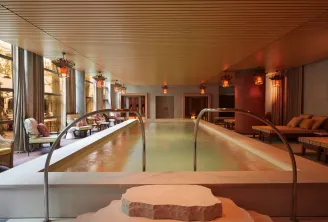 Luxurious indoor spa featuring two thermal pools, light wooden ceiling, warm ambient lighting, contemporary furnishings, and floor-to-ceiling windows overlooking Bordeaux