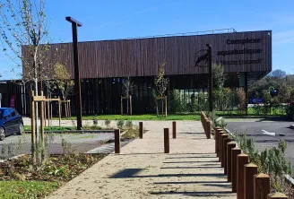 Entrance to the Elodie Lorandi water complex in Bordeaux featuring a modern wooden and steel building, paved pathway with bollards, trees and green spaces