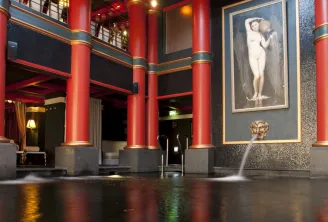 Interior of Guerlain Spa in Bordeaux featuring red ornamental columns, reflective floor, central fountain, and classical framed portrait on textured gray wall.