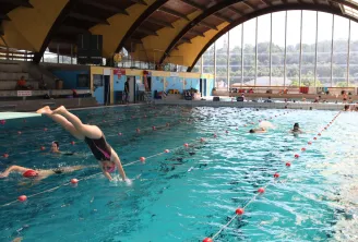 Interior view of Bordeaux's municipal swimming pool Galin with multiple swimmers and a diver in mid-dive, under a yellow vaulted roof and large windows overlooking the city