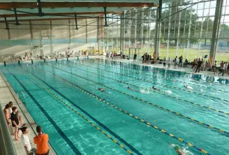 Interior view of the Olympic swimming pool of Villenave d'Ornon in Bordeaux with multiple swimmers in the lanes, spectators in the stands, and a large covered facility with metal structure.