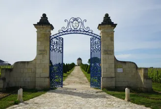 Ornate blue and gold entrance gate of Château Balestard La Tonnelle in Bordeaux, flanked by two stone pillars, leading to a cypress-lined avenue towards the château