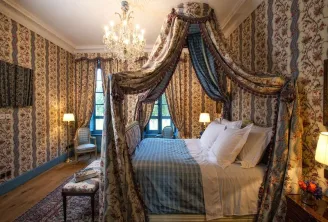 Elegant bedroom at Château Soutard in Bordeaux featuring a canopy bed with ornate draped fabrics, floral patterned wallpaper, crystal chandelier, and classic furnishings.