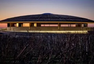 The Dome of Bordeaux glowing at dusk, a modern rounded building in a harvested cornfield with lit windows against a blue and orange twilight sky.