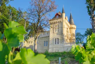 Château de Roques near Bordeaux, a white stone neo-gothic castle with two pointed towers, surrounded by vineyards and trees under a clear blue sky.