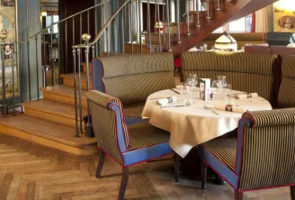Elegant dining room of Le Bordeaux restaurant featuring striped blue and gold armchairs, a set round table, wooden staircase, and historical displays on the walls.