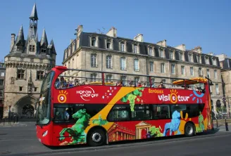 A red double-decker sightseeing bus from Bordeaux Visiotour (Hop On Hop Off) service parked in front of a medieval tower and Haussmann-style buildings in Bordeaux