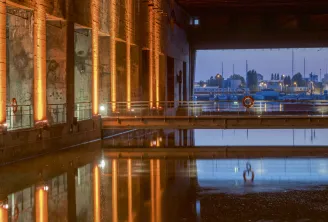 Night view of Bordeaux's submarine base featuring illuminated orange columns reflected in still water, overlooking the port and Garonne river at dusk.
