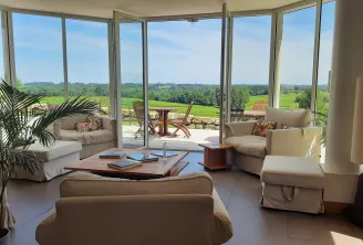 Bright living room with large glass doors overlooking Bordeaux vineyards, elegant beige furniture, wooden coffee table, outdoor terrace and panoramic views of green fields at Logis de Valandraud.