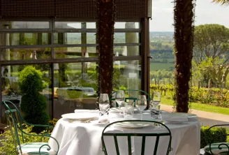 Fine dining table set with white tablecloth, glassware, and place settings on a terrace overlooking Bordeaux vineyards and valley landscape, framed by stone columns and large windows.